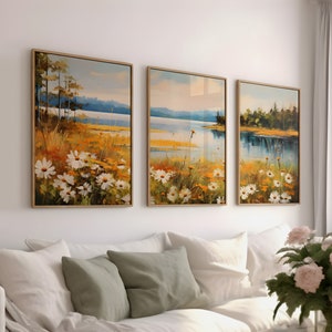 May include: Three framed landscape paintings depicting a lake, trees, and white flowers, displayed in a living room with a white sofa and a coffee table.