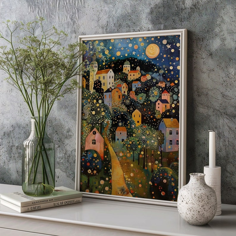 May include: A framed art print depicting a whimsical village scene under a starry night sky. The artwork features colorful houses, trees, and a winding path, with a large moon and stars. The print is displayed on a white surface with a vase of greenery and a candle.