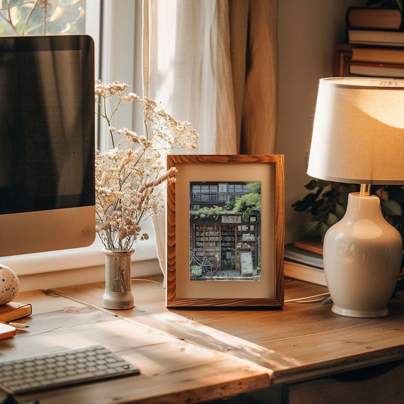 May include: A wooden picture frame with a photo of a storefront with a bicycle parked in front. The photo is in a vintage style and the storefront has a sign that says "Bookstore". The frame is sitting on a wooden table with a lamp and a vase of flowers.