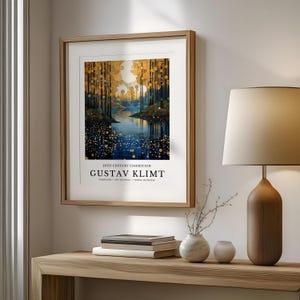 May include: A framed art print featuring a landscape painting with a river and trees in a forest. The artwork has a gold and blue color palette and includes the text "20TH CENTURY EXHIBITION GUSTAV KLIMT". The print is displayed on a wooden table.