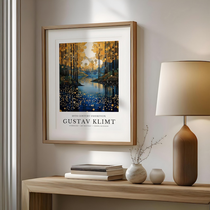 May include: A framed art print featuring a landscape painting with a river and trees in a forest. The artwork has a gold and blue color palette and includes the text "20TH CENTURY EXHIBITION GUSTAV KLIMT". The print is displayed on a wooden table.