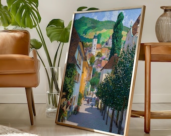 May include: A rectangular framed wall art print featuring a vibrant oil painting of a European town street. The scene depicts cobblestone paths, colorful buildings in shades of yellow and blue, lush green trees, and a distant church tower under a blue sky.