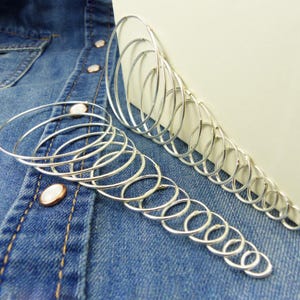 May include: Silver hoop earrings of varying sizes, arranged in a cascading pattern. The earrings are displayed on a denim shirt, highlighting their reflective surface. The hoops' diameters vary, creating a layered effect.