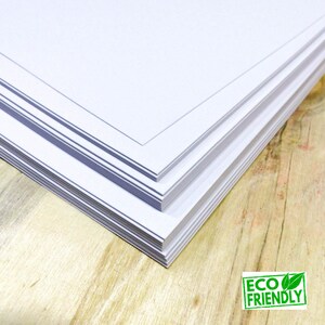 May include: A stack of white paper with a black border on the top sheet. The paper is on a light brown wooden surface. The image has a green "ECO FRIENDLY" label with a leaf design.