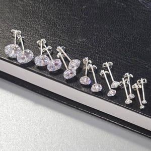 May include: A selection of silver stud earrings, each featuring a clear, oval-cut cubic zirconia. The earrings are displayed on a dark surface, highlighting their sparkle. The earrings vary in size and design, showcasing different styles.