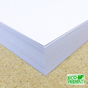 May include: A stack of white paper, likely printer paper, with a corner folded over. The paper is on a brown surface. The text "ECO FRIENDLY" is visible in green letters on a green leaf background.