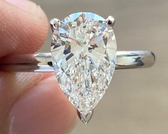 Puede incluir: Un anillo de compromiso de diamante de forma pera engastado en una banda plateada. El diamante es claro y brillante.