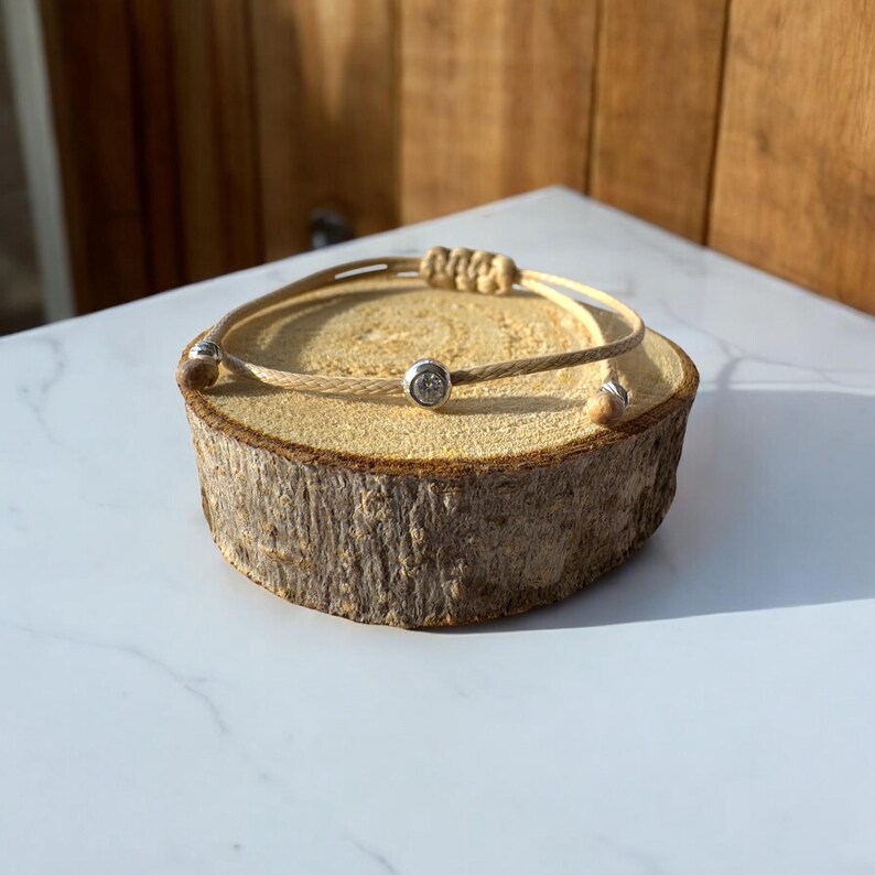 May include: A simple beige cord bracelet with two silver beads. The bracelet is displayed on a wooden log.