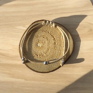 May include: A simple beige rope bracelet with silver beads. The bracelet is displayed on a wooden surface.