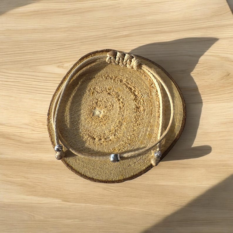 May include: A simple beige rope bracelet with silver beads. The bracelet is displayed on a wooden surface.