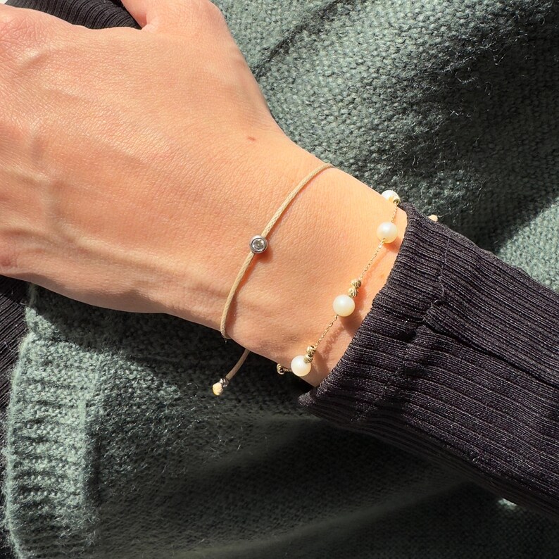 May include: Two bracelets on a wrist. One bracelet is a simple beige cord with a small clear gemstone. The other bracelet is a gold chain with small white pearls.