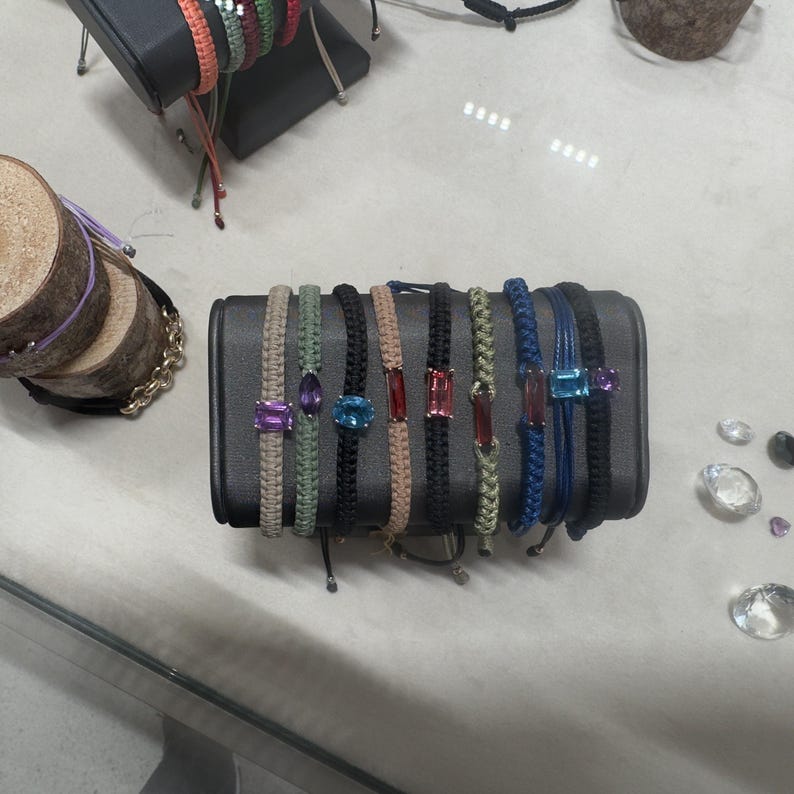 May include: A display of colorful braided bracelets with gemstone accents. The bracelets are arranged in a row on a black surface. The gemstones include amethyst, turquoise, and ruby.