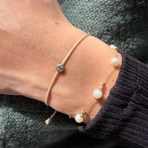 May include: Two bracelets on a wrist. One bracelet is a simple beige string with a small diamond charm. The other bracelet is a gold chain with white pearls.