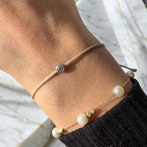 May include: Two bracelets on a wrist. The top bracelet is a simple beige cord with a small diamond accent. The bottom bracelet is a delicate gold chain with white pearls and gold accents.