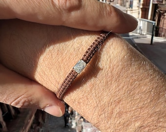 May include: A brown braided bracelet with a small, square-shaped, clear stone accent.