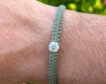 May include: A light green braided bracelet with a single, clear, round-cut gemstone. The bracelet is centered on the wrist, showcasing the sparkling stone. The background features blurred green foliage and pink flowers.