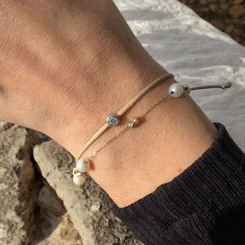May include: Two bracelets on a wrist. The top bracelet is a beige cord with a small pearl charm. The bottom bracelet is a gold chain with a small diamond charm and two pearl charms.