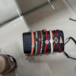 May include: A set of eight colorful braided bracelets with different charms, including a round diamond, a teardrop diamond, and a triangle charm. The bracelets are displayed on a black stand.