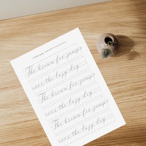 K&ouml;nnte beinhalten: Ein Kalligraphie-Arbeitsblatt mit grauen Linien und dem Text "The brown fox jumps over the lazy dog", der dreimal wiederholt wird.