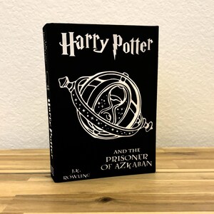 May include: A black book with a silver title, "Harry Potter and the Prisoner of Azkaban" by J.K. Rowling. The cover features a silver illustration of a time-turner.