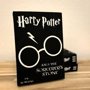 Könnte beinhalten: Ein schwarzes Buch mit dem Titel "Harry Potter und der Stein der Weisen" von J.K. Rowling. Der Buchumschlag zeigt einen weißen Blitz und eine Brille.
