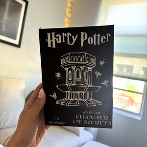 May include: A black book cover with white text that reads "Harry Potter and the Chamber of Secrets" by J.K. Rowling. The cover features a white illustration of a tower with a pointed roof and a series of arches.