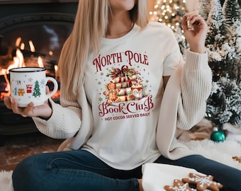 Puede incluir: Camiseta color crema con un gráfico festivo de "North Pole Book Club", con libros apilados, hombres de jengibre y una taza de chocolate caliente. Se sostiene una taza blanca con diseños navideños.