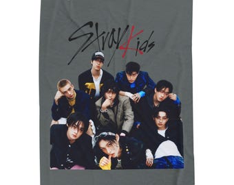 Puede incluir: Una manta gris con un grupo de nueve personas. La manta tiene las palabras "Stray Kids" en escritura negra y roja en la parte superior. Las personas visten varios atuendos, incluyendo chaquetas, camisas y sombreros.
