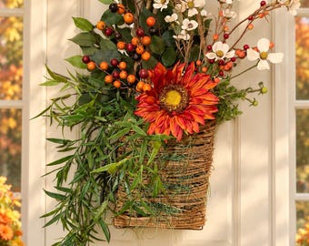 May include: A hanging wall basket arrangement featuring a large orange sunflower, white cosmos flowers, green foliage, and clusters of orange and dark red berries. The basket is constructed from woven brown twigs and hangs from a jute rope loop.