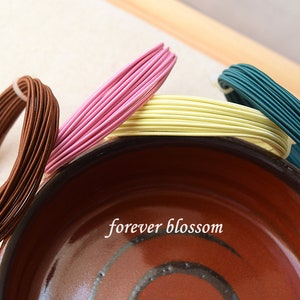 May include: An assortment of colorful wire bundles in brown, pink, yellow, and teal, arranged near a brown ceramic bowl. The text "forever blossom" is visible on the bowl. These wires are likely for crafting or jewelry making.