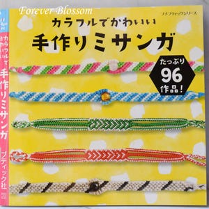 Może przedstawiać: Okładka książki z tytułem "Forever Blossom" i japońskim tekstem, przedstawiająca kolorowe, ręcznie robione bransoletki przyjaźni. Bransoletki są w różnych wzorach i kolorach, w tym różowym, zielonym, niebieskim i pomarańczowym. Czarny sześciokąt wyświetla "96 prac!"