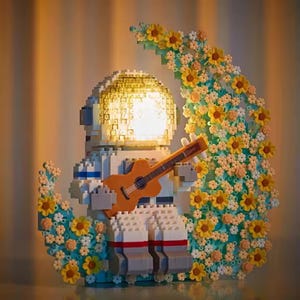 Könnte beinhalten: Eine weiße Astronautenfigur, die Gitarre spielt, aus Bausteinen gefertigt, mit einem Licht im Helm. Der Astronaut sitzt auf einem Halbmond aus gelben und blauen Blumen.