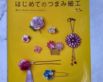 Puede incluir: Un libro instructivo amarillo titulado "Hajimete no Tsumami Saiku" que muestra varias manualidades de flores de tela en la portada, incluyendo horquillas, un broche y un llavero con pétalos morados, rosas, azules y blancos y pequeñas cuentas.