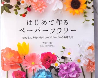 May include: A book cover titled "はじめて作るペーパーフラワー" featuring a vibrant arrangement of handcrafted paper flowers including red roses, a yellow sunflower, pink dahlias, blue hydrangeas, and green stems against a white background.