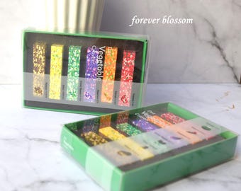 May include: Two rectangular green boxes containing sets of Vegetable Crayon sticks. Each box holds six multicolored, speckled crayons labeled with vegetable names like Potato, Corn, Green Pepper, Eggplant, Carrot, and Tomato in white text.