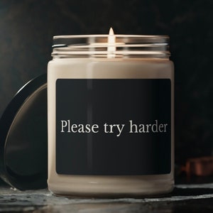 May include: A glass candle jar with a black label that reads "Please try harder". The candle is lit and the wax is a light beige color.