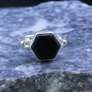 Op de afbeelding: Een zilveren ring met een zeshoekige zwarte steen. De ring heeft een eenvoudige band met kleine, ronde versieringen aan weerszijden van de steen. De zwarte steen is het middelpunt, gezet in een zilveren zetting.
