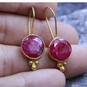 May include: A pair of gold-colored drop earrings. Each earring features a large, round, faceted, deep pink gemstone set in a gold bezel. Below the gemstone, three small gold beads add detail. The earrings have a simple hook design.