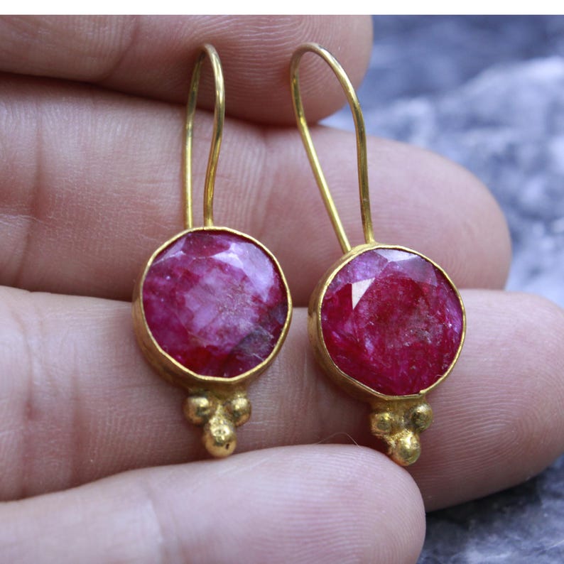 May include: A pair of gold-colored drop earrings. Each earring features a large, round, faceted, deep pink gemstone set in a gold bezel. Below the gemstone, three small gold beads add detail. The earrings have a simple hook design.