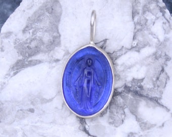 May include: A silver pendant with a blue glass cameo of the Virgin Mary. The cameo is surrounded by a silver frame and has a silver bail.