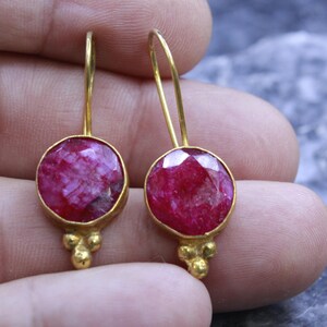 May include: A pair of gold-colored drop earrings. Each earring features a round, faceted, ruby-red gemstone set in a gold bezel. The earrings have a simple hook design and a cluster of three small gold beads at the bottom.
