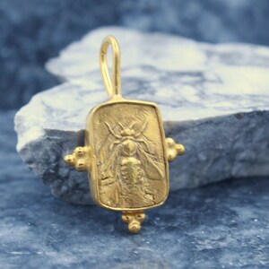 May include: Gold pendant featuring a detailed bee design. The pendant is rectangular with a textured surface and has a loop at the top for attaching to a chain.