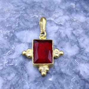 May include: A gold-toned pendant featuring a rectangular, faceted deep red stone set in a metal frame. The pendant has a small loop at the top for a chain and decorative beaded metal accents on the left, right, and bottom sides of the stone.