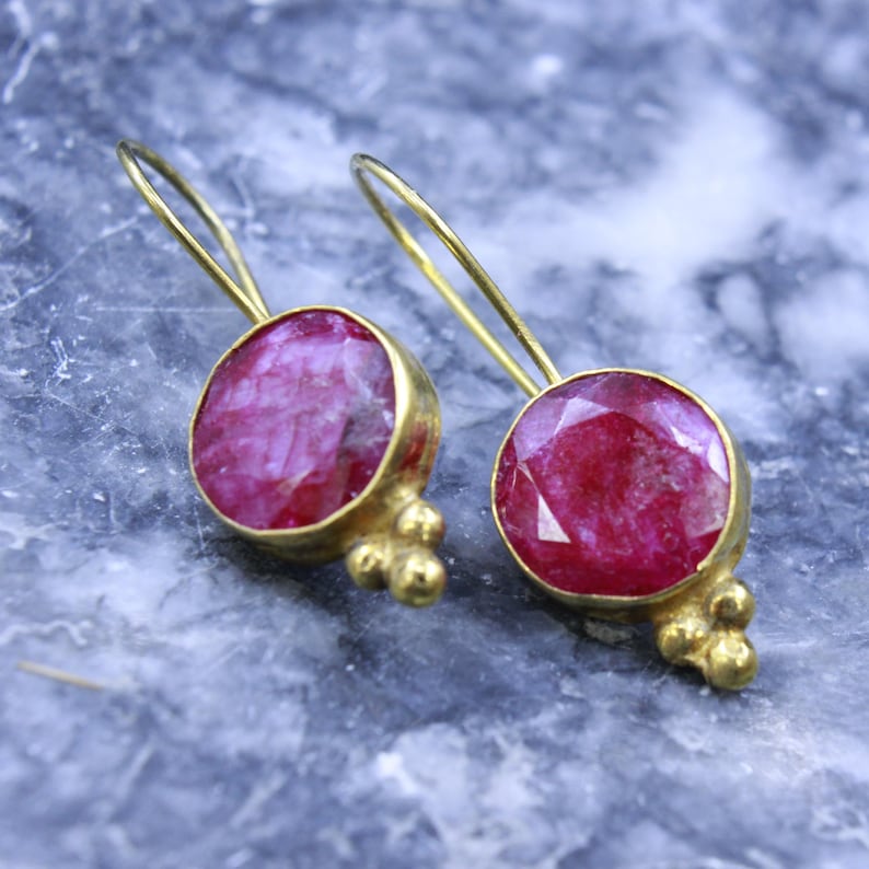 May include: A pair of gold-colored drop earrings featuring large, round, faceted, ruby-red gemstones. The earrings have a simple design with a hook and a cluster of small gold beads at the base of the gemstone setting.