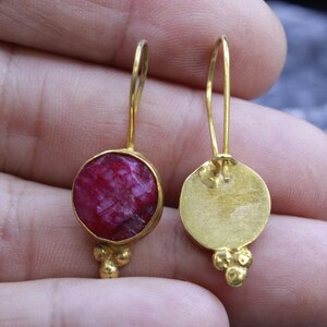 May include: A pair of gold-colored earrings. Each earring features a round, deep red gemstone set in a gold bezel. The earrings have a simple hook design and a cluster of three small gold beads at the bottom.