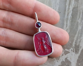 May include: A red glass pendant with a silver setting and a carved insect design. The pendant has a small red gemstone at the top.