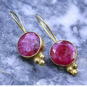 May include: A pair of gold-colored drop earrings featuring large, round, faceted, ruby-red gemstones. The earrings have a simple, elegant design with a small cluster of gold beads at the base of each gemstone.