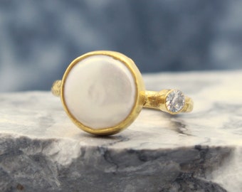 May include: A gold ring with a white pearl and a small clear stone accent. The ring is on a gray textured surface.