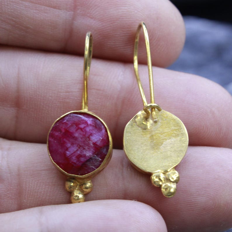 May include: A pair of gold-colored earrings. Each earring features a round, faceted, deep red gemstone set in a gold bezel. The earrings have a simple hook design and a cluster of three small gold beads at the bottom.