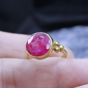 May include: A gold ring featuring a large, round, faceted ruby gemstone. The ruby is a vibrant shade of red. The ring has a simple, elegant design with a cluster of three small gold beads on the side.